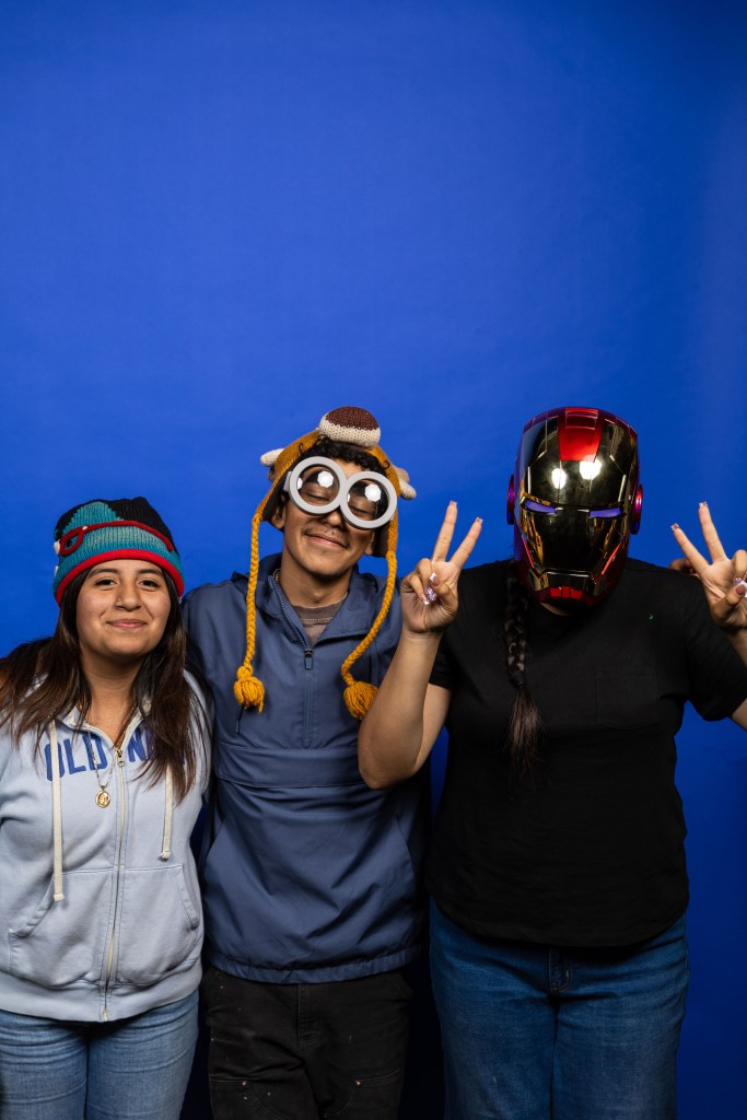 Three friends posing against a blue background, wearing playful hats and accessories. The person on the left wears a colorful beanie, the middle person wears a whimsical hat with goggles, and the person on the right sports an Iron Man helmet. All are smiling and making peace signs.