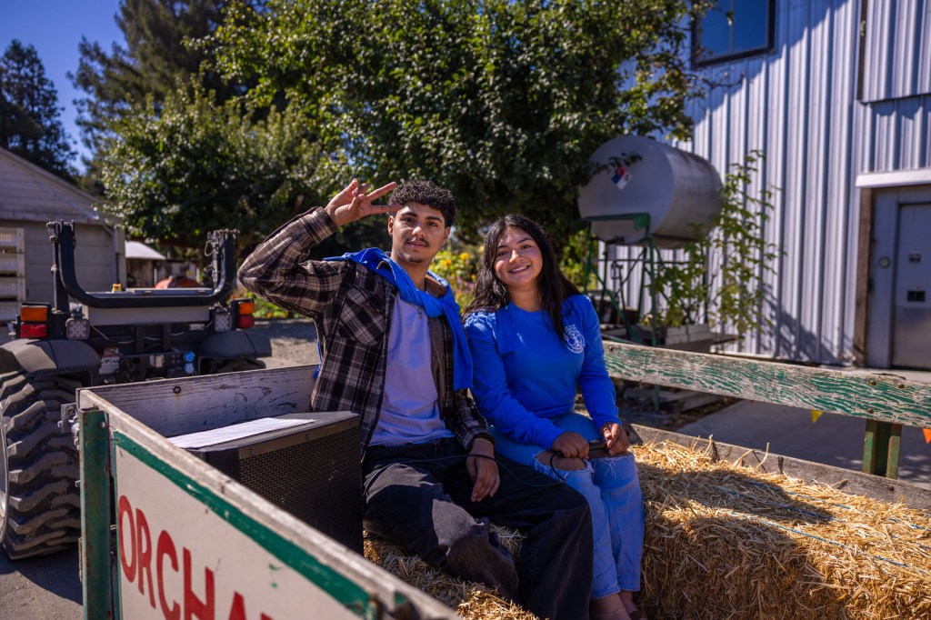 Two young adults sitting on a hay-filled wagon in a farm setting, smiling at the camera with a tractor in the background.