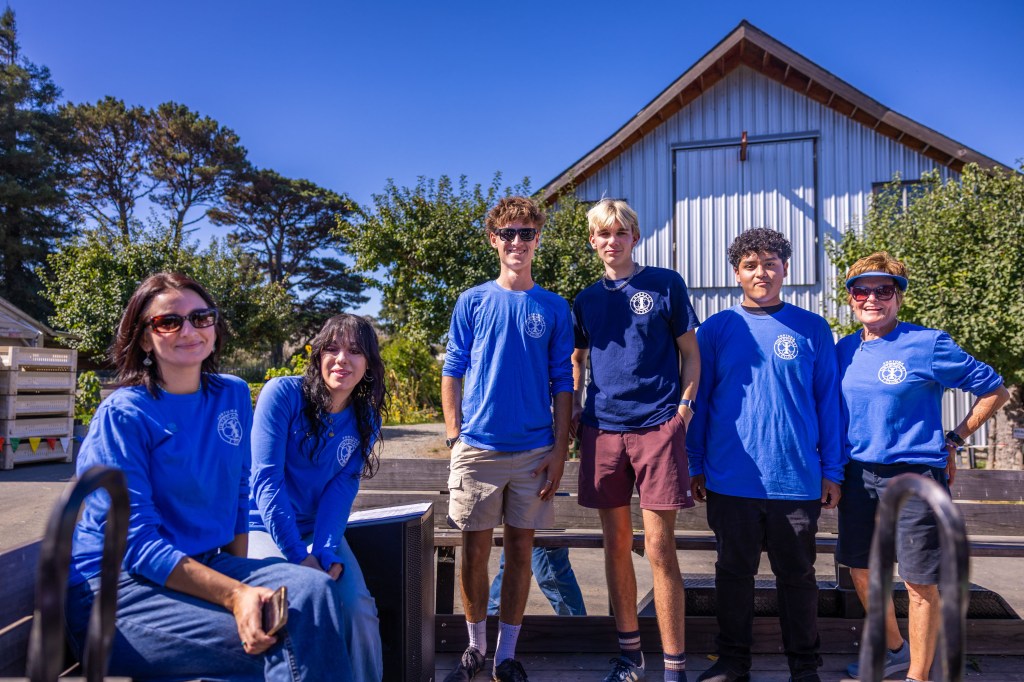 A group of six people smiling and posing outdoors, wearing blue shirts, with trees and a building in the background.