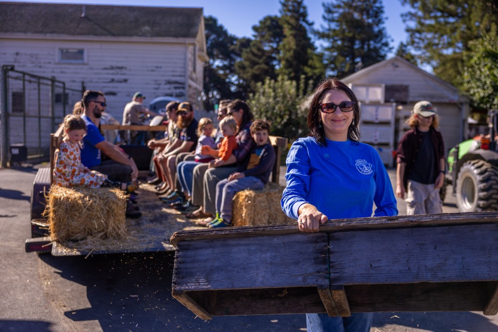 A woman in a blue shirt smiles while holding a wooden board, with a group of people sitting on hay bales in the background. It is a sunny day and there are trees and buildings nearby.