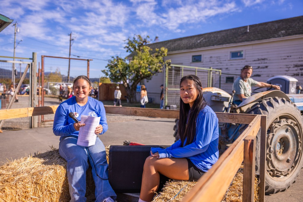 Two girls sitting on a hay bale in a wooden trailer, one holding a microphone and a paper, with a tractor and more people in the background under a blue sky.