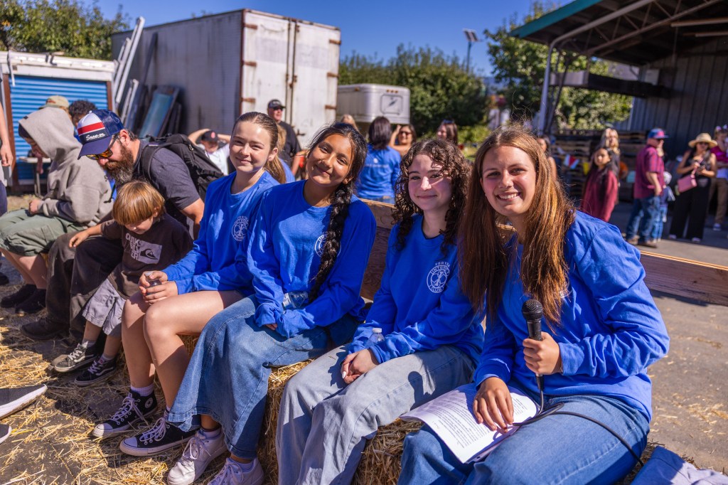 A group of four young girls wearing blue long-sleeve shirts sit on a hay bale at an event, smiling and posing for the camera. One of the girls holds a microphone, while others have notes in hand. People are gathered in the background.